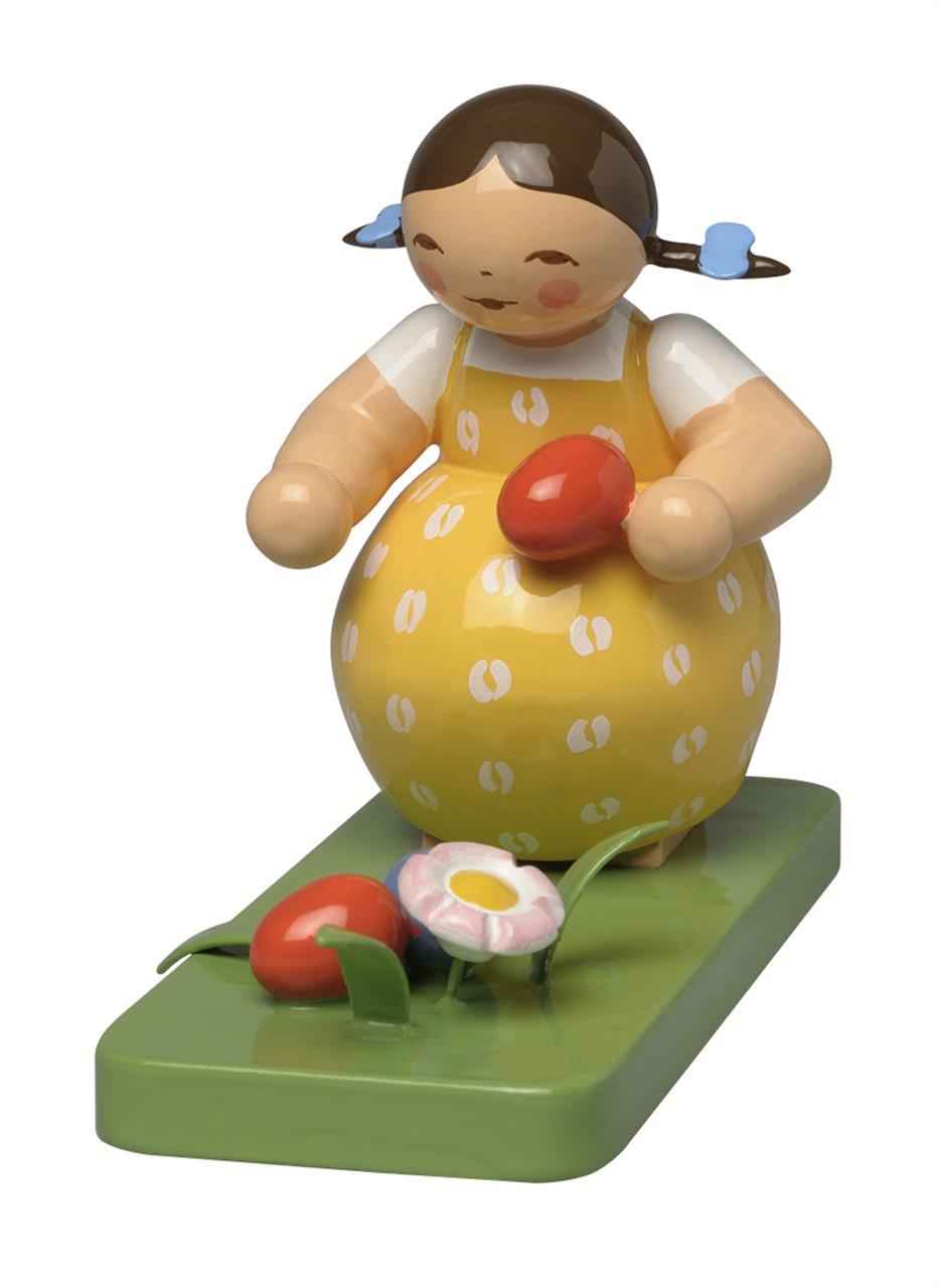 Das Bild zeigt eine bunte Holzfigur eines Mädchens im gelben Kleid mit roten Ostereiern. Die Figur steht auf einer grünen Platte mit Blumen und Ostereiern.