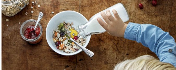 Ein Kind gießt Milch in eine Schüssel mit Müsli und Obst. Daneben steht ein Glas mit Marmelade auf einem Holztisch.