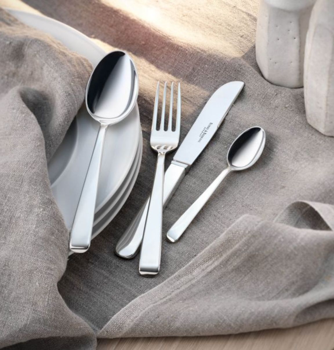 Auf dem Bild liegt silbernes Besteck bestehend aus einem Löffel, einer Gabel, einem Messer und einem kleinen Löffel auf einem grauen Tuch. Darunter sind weiße Teller zu sehen.