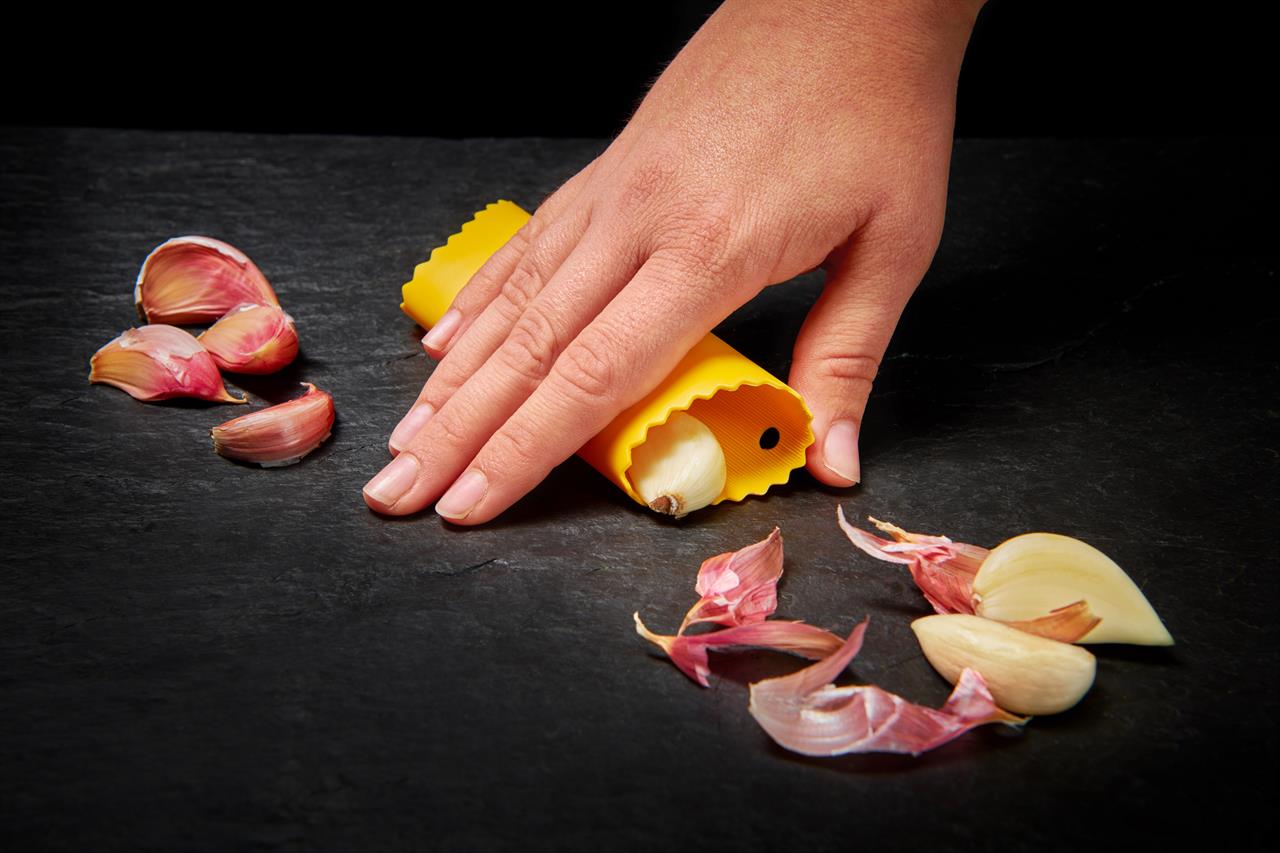 Eine Hand benutzt einen gelben Knoblauchschäler, um eine Knoblauchzehe zu schälen. Um den Schäler herum liegen bereits geschälte und ungeschälte Knoblauchzehen sowie Knoblauchschalen.