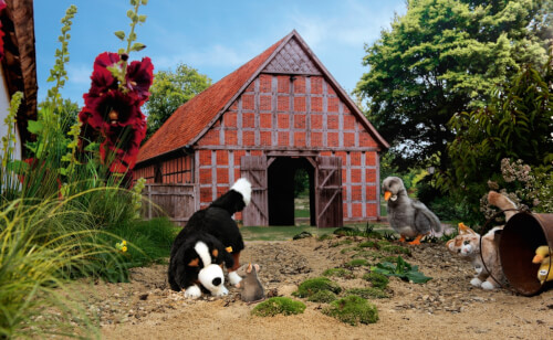 Im Vordergrund spielen mehrere Stofftiere, darunter ein Hund und ein kleines Mäuschen, auf einem Sandweg. Im Hintergrund steht eine Scheune mit roten Dachziegeln, umgeben von Bäumen und Blumen.