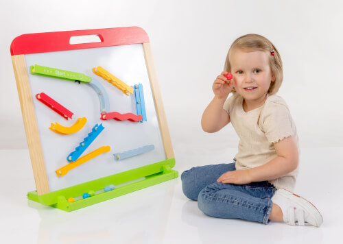 Ein kleines Kind sitzt auf dem Boden und spielt mit einer Kugelbahn aus Holz. Das Kind hält eine rote Kugel in der Hand und lächelt in die Kamera.