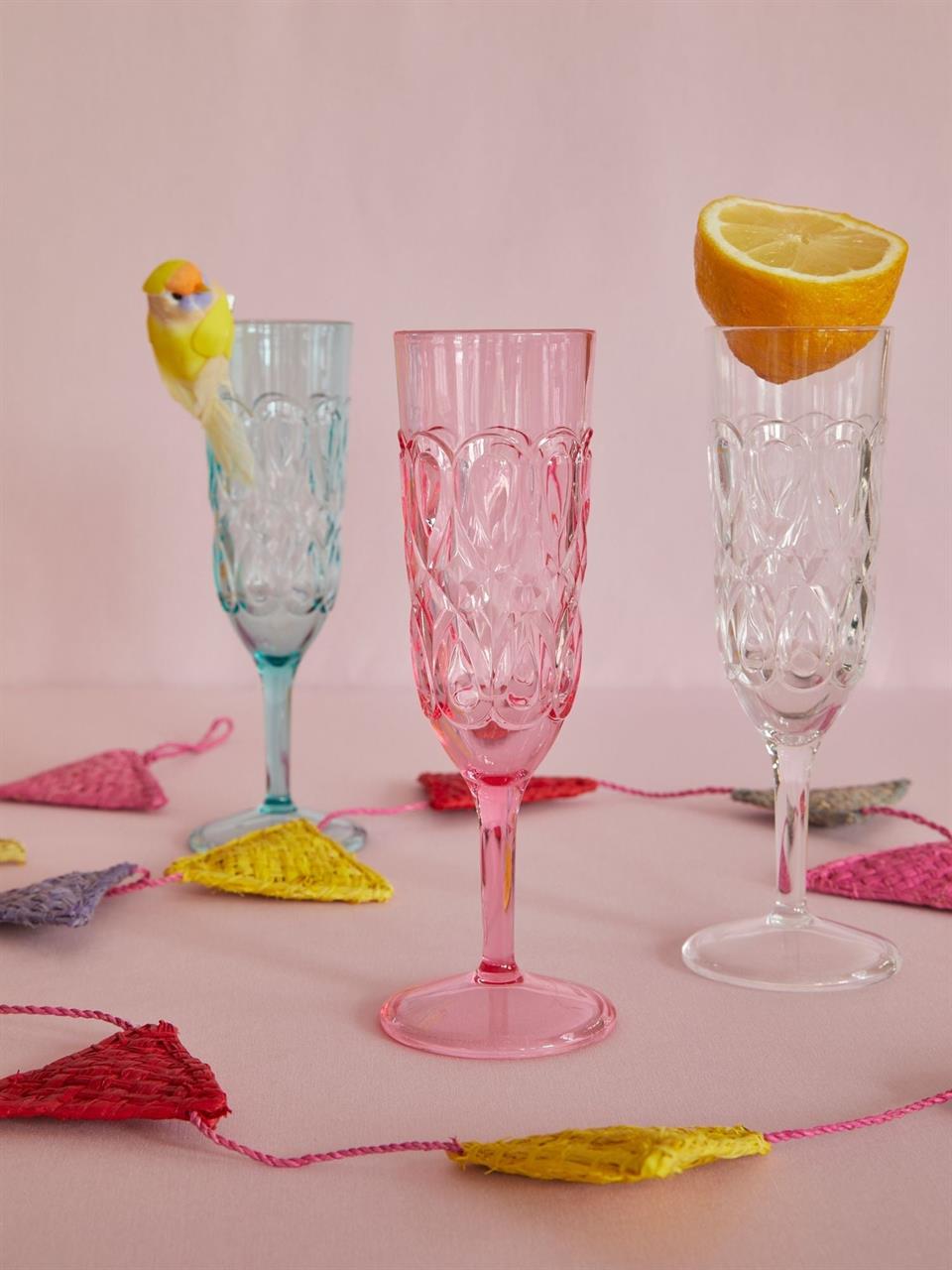 Auf dem Bild sind drei Sektkelche aus Acryl mit geschwungenem Prägung zu sehen, einer davon in Mintfarbe. Einer der Gläser ist mit einem künstlichen Vogel dekoriert, ein anderer mit einer Orangenscheibe, und bunte Girlanden liegen auf dem Tisch.