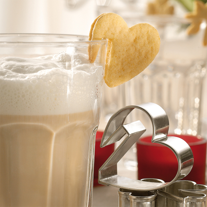 Ein Glas mit Milchschaum steht auf dem Tisch, darauf steckt ein Keks in Herzform. Daneben liegt ein herzförmiger Ausstecher, der das Ausstechen der Plätzchen ermöglicht.