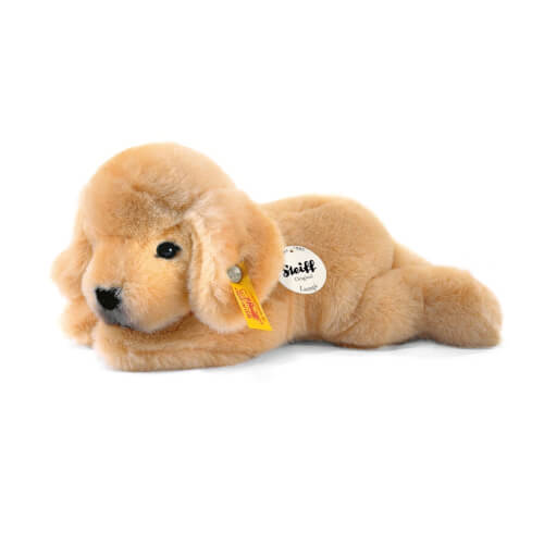 Das Bild zeigt einen hellbraunen Plüschhund in liegender Position. Am Ohr trägt er ein gelbes Etikett und an der Seite ein rundes weißes Schild mit der Aufschrift „Steiff“.