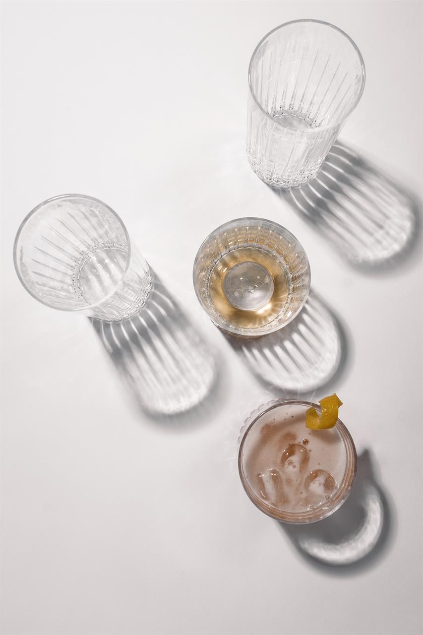 Das Bild zeigt vier stilvolle Longdrinkgläser auf einer weißen Oberfläche. Zwei Gläser sind leer, eines enthält ein Getränk mit Eis und Zitronenzeste, und ein weiteres ist mit einer goldenen Flüssigkeit gefüllt.