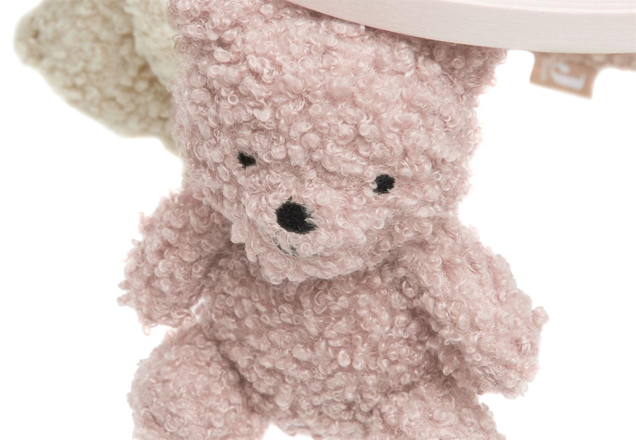 Das Bild zeigt einen kuscheligen Teddybär in der Farbe Wild Rose, der an einem Baby-Mobile befestigt ist. Im Hintergrund ist ein weiterer Teddybär in Naturfarbe zu sehen.