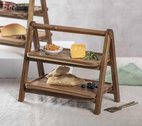 Auf dem Bild ist ein zweistöckiger Servierturm aus Akazienholz mit dem Namen „Gather“ zu sehen. Auf den Etagen liegen verschiedene Speisen wie Brot, Käse und Beeren.