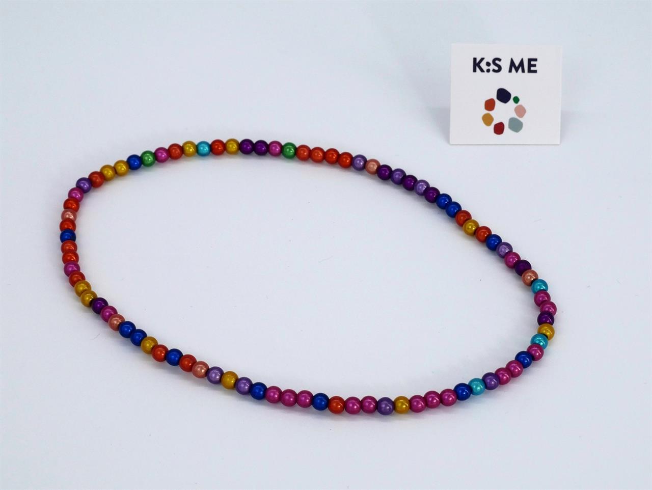 Die Abbildung zeigt eine Kette mit kleinen, bunten Perlen in dunklen Farben. Im Hintergrund ist ein Schild mit der Aufschrift "K:S ME" zu sehen.