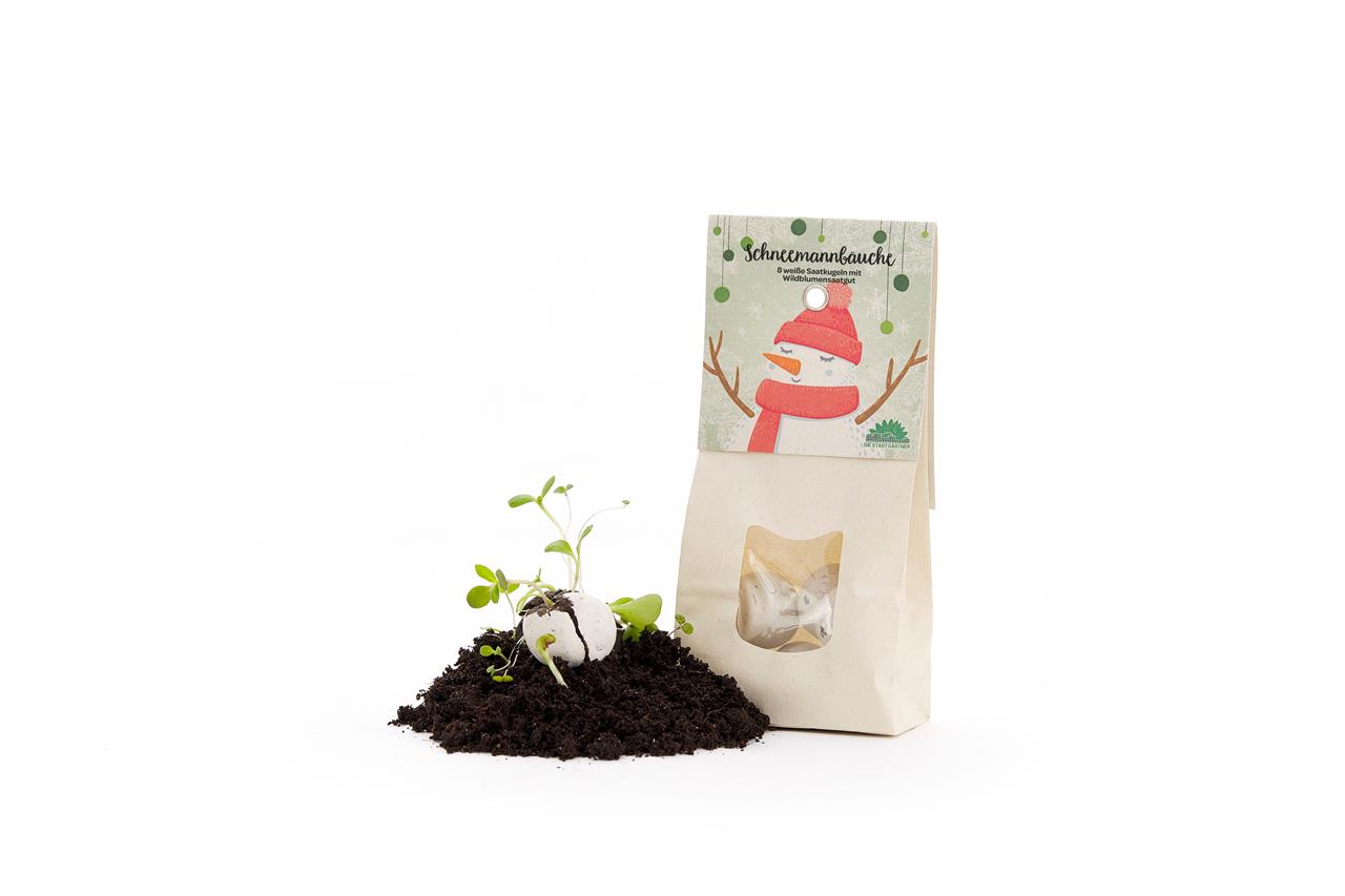 Das Bild zeigt eine Packung "Schneemannbäuche - Weiße Blumenmurmeln" neben etwas Erde und einer keimenden Samenmurmeln. Auf der Verpackung ist ein Schneemann mit rotem Schal und Mütze abgebildet.