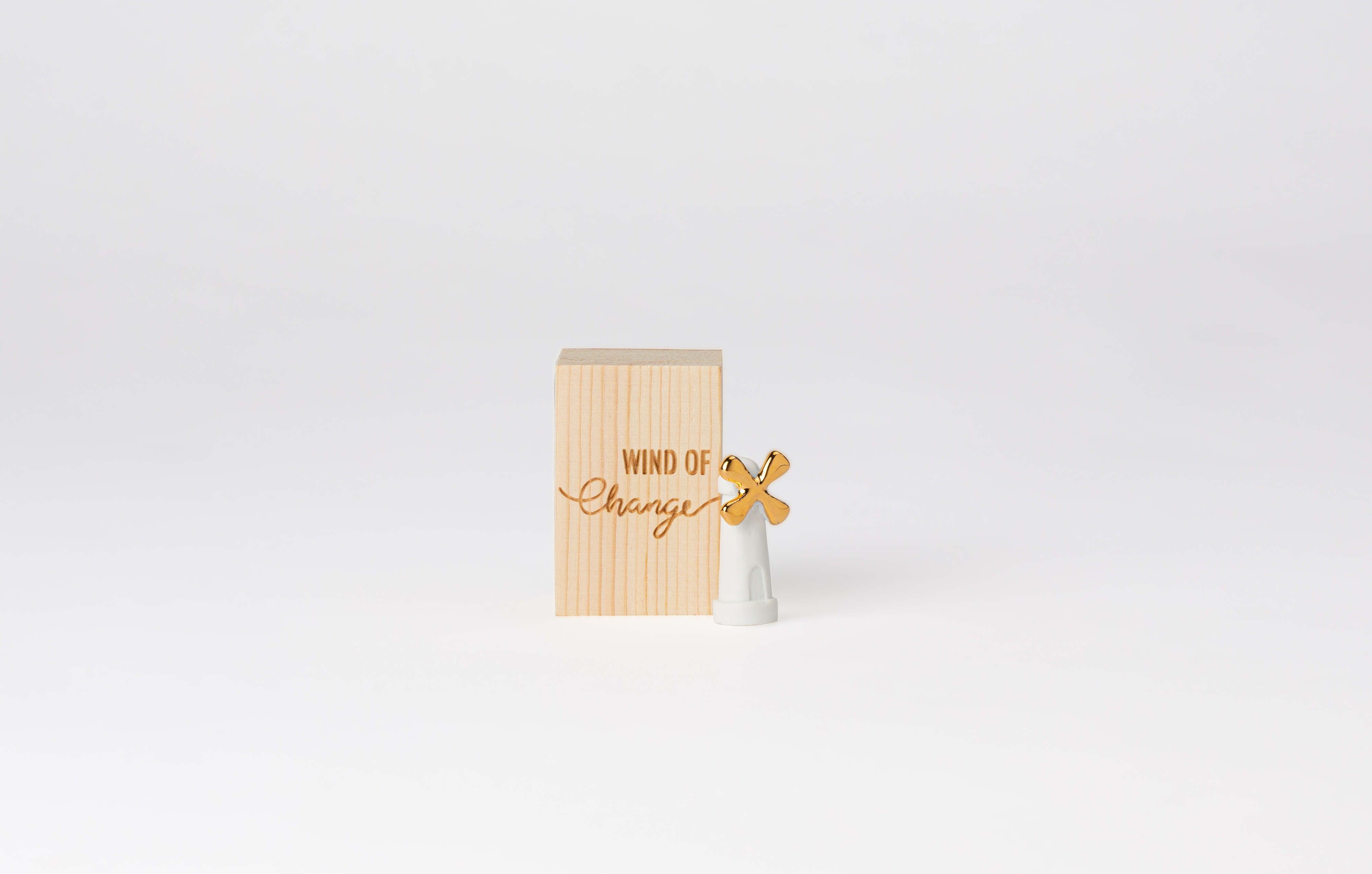 Auf dem Bild ist eine kleine Holzbox mit der Aufschrift "WIND OF Change" zu sehen. Daneben steht eine kleine weiße Figur mit einem goldenen Windrad.