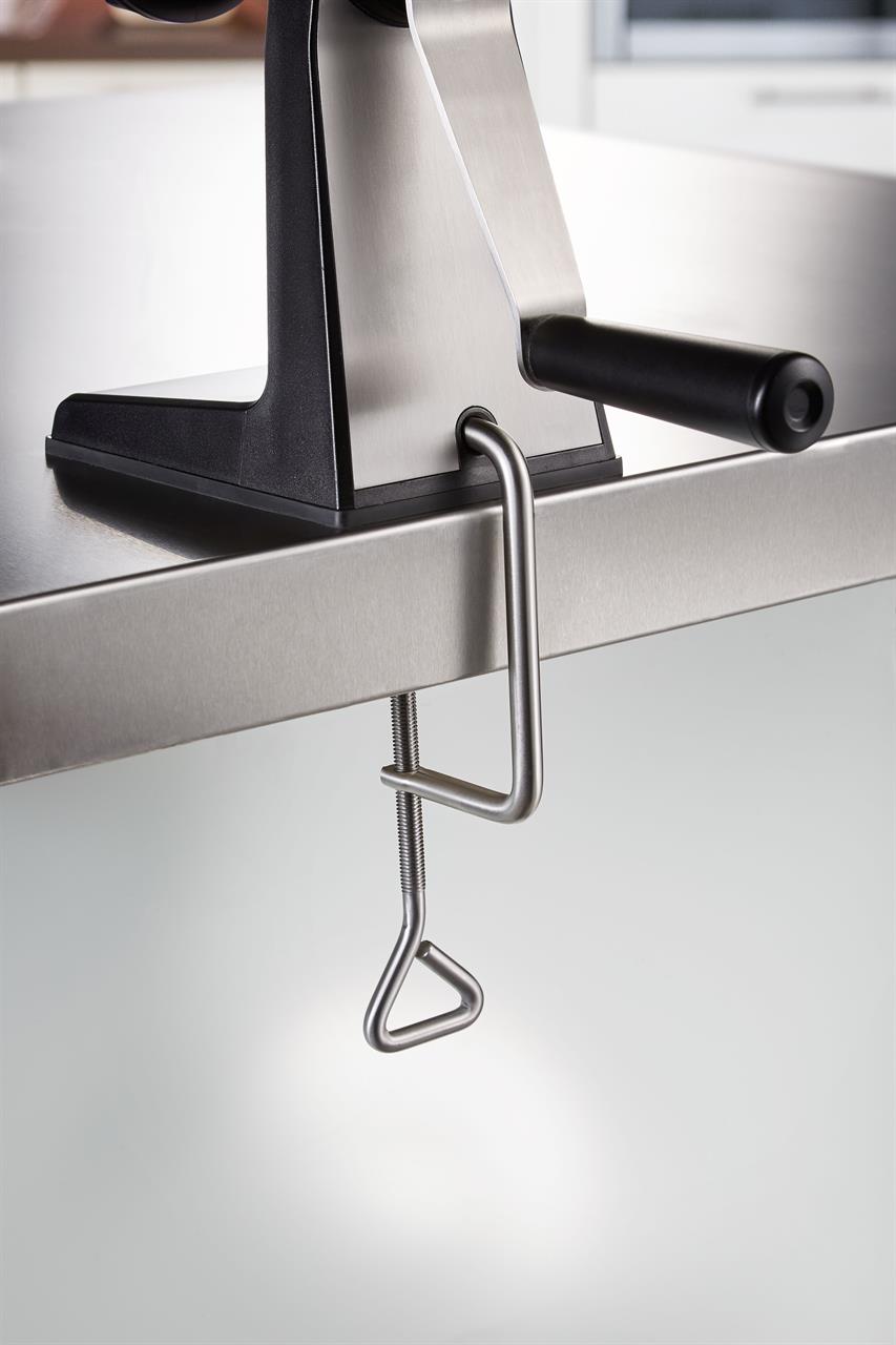 Das Bild zeigt einen Fleischwolf Transforma, der mit einer Klemme an einer Tischkante befestigt ist. Ein schwarzer Griff ist an der Seite zum Drehen angebracht.