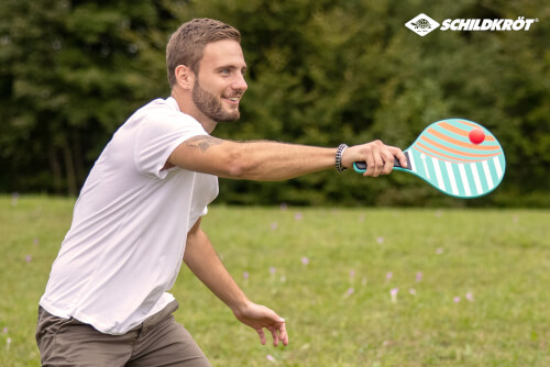 Ein Mann spielt mit einem bunten Schläger und einem roten Ball im Freien. Im Hintergrund ist eine grüne Wiese und Bäume zu sehen.