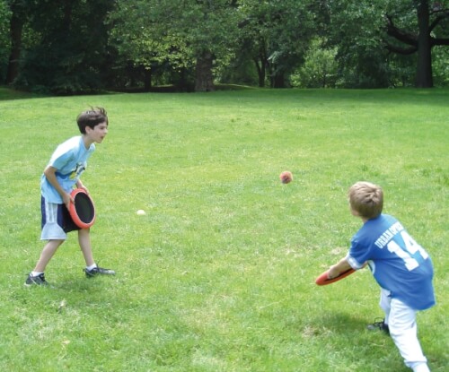 Zwei Jungen spielen auf einer Wiese mit Fangscheiben und einem Ball. Einer der Jungen wirft den Ball, während der andere bereit ist, ihn zu fangen.
