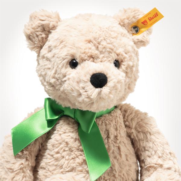 Der Teddybär Jimmy 30 Beige Good Luck ist ein weicher, beigefarbener Teddybär mit einem grünen Band um den Hals. Er hat schwarze Knopfaugen und trägt ein gelbes Steiff-Schild am Ohr.