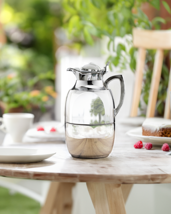 Auf dem Bild steht eine silberne Thermoskanne auf einem Holztisch. Im Hintergrund sind Geschirr, Kuchen und frische Himbeeren zu sehen, die eine Gartenatmosphäre schaffen.