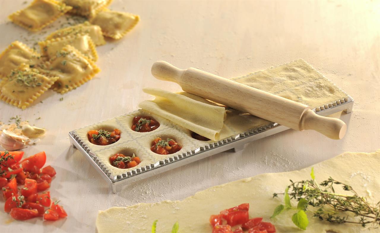Das Bild zeigt ein Ravioli/Maultaschenbrett mit Teig und Füllung, bereit zum Zubereiten. Ein Nudelholz liegt auf dem Brett, und im Hintergrund sind bereits fertige Ravioli zu sehen.