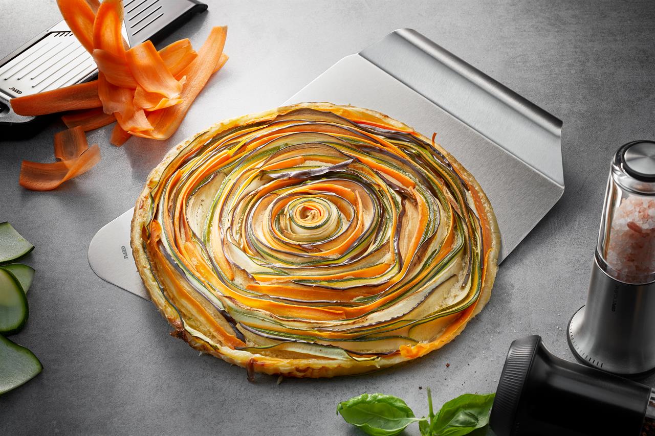 Auf dem Bild ist ein Kuchenheber namens "Kuchen- u. Pizzaheber Easy" zu sehen. Mit ihm wird eine bunte Gemüsetarte auf einer Arbeitsfläche serviert.