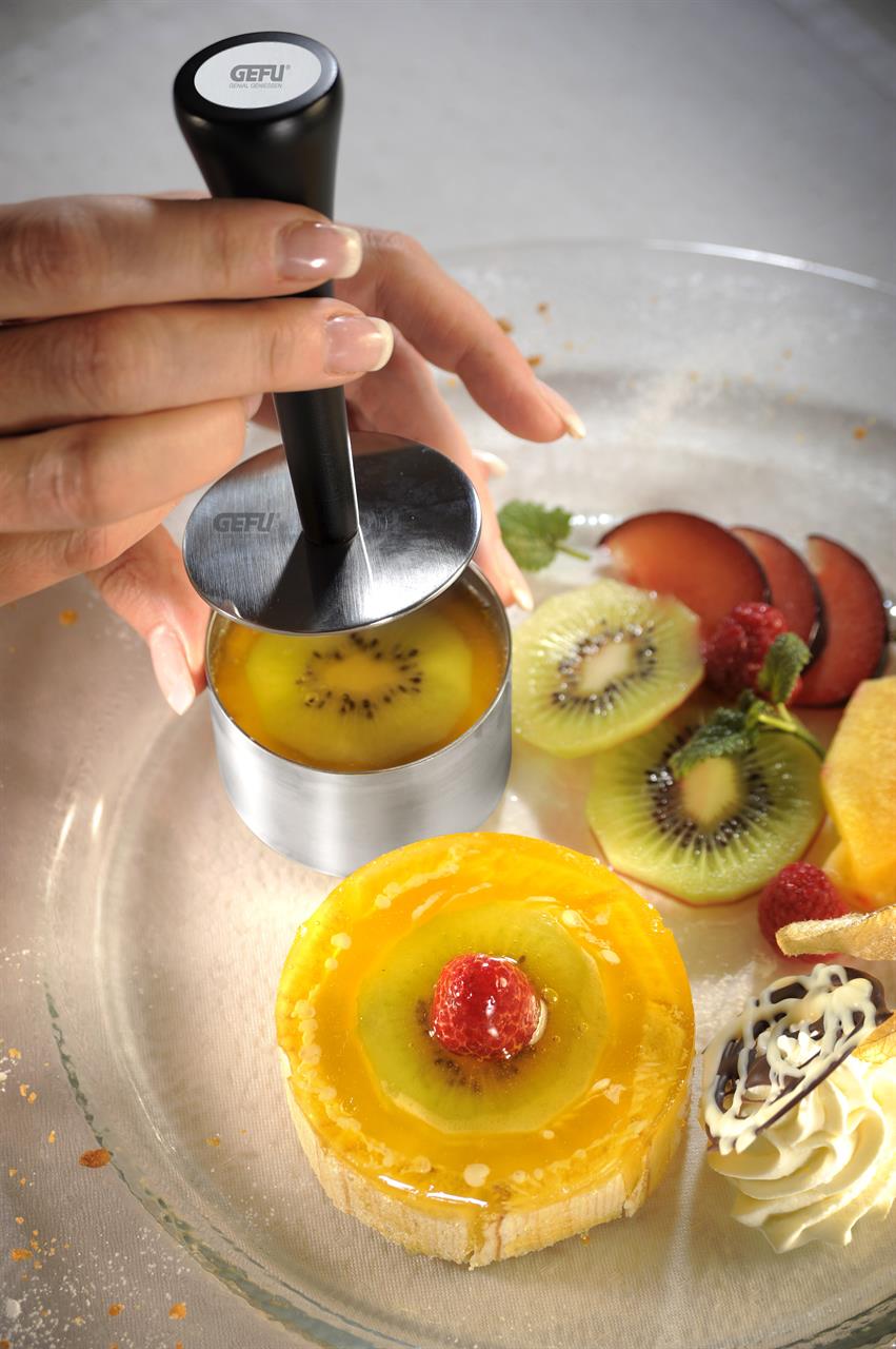 Das Bild zeigt eine Hand, die einen Profispeisenring aus dem Set "Formidable, 10-tlg." benutzt, um ein Dessert in Form zu bringen. Frische Früchte und ein fertig angerichtetes Dessert sind ebenfalls auf dem Teller zu sehen.