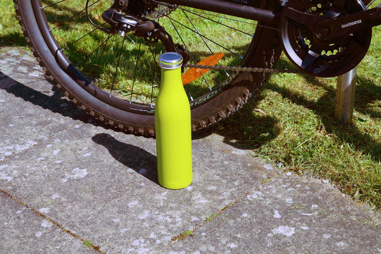 Eine Isolierflasche aus Edelstahl in der Farbe Fresh Green steht auf dem Gehweg. Im Hintergrund ist das Hinterrad eines Fahrrads zu sehen.