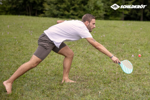Ein Mann spielt draußen auf einer Wiese mit einem bunten Schläger und einem Ball. Er ist barfuß und streckt sich, um den Ball zu erreichen.