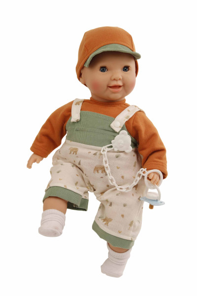Die Puppe trägt eine braune Mütze, ein orangefarbenes Oberteil und eine Latzhose mit Waldmotiv. Sie hat blaue Schlafaugen und hält einen Schnuller an einer Kette.