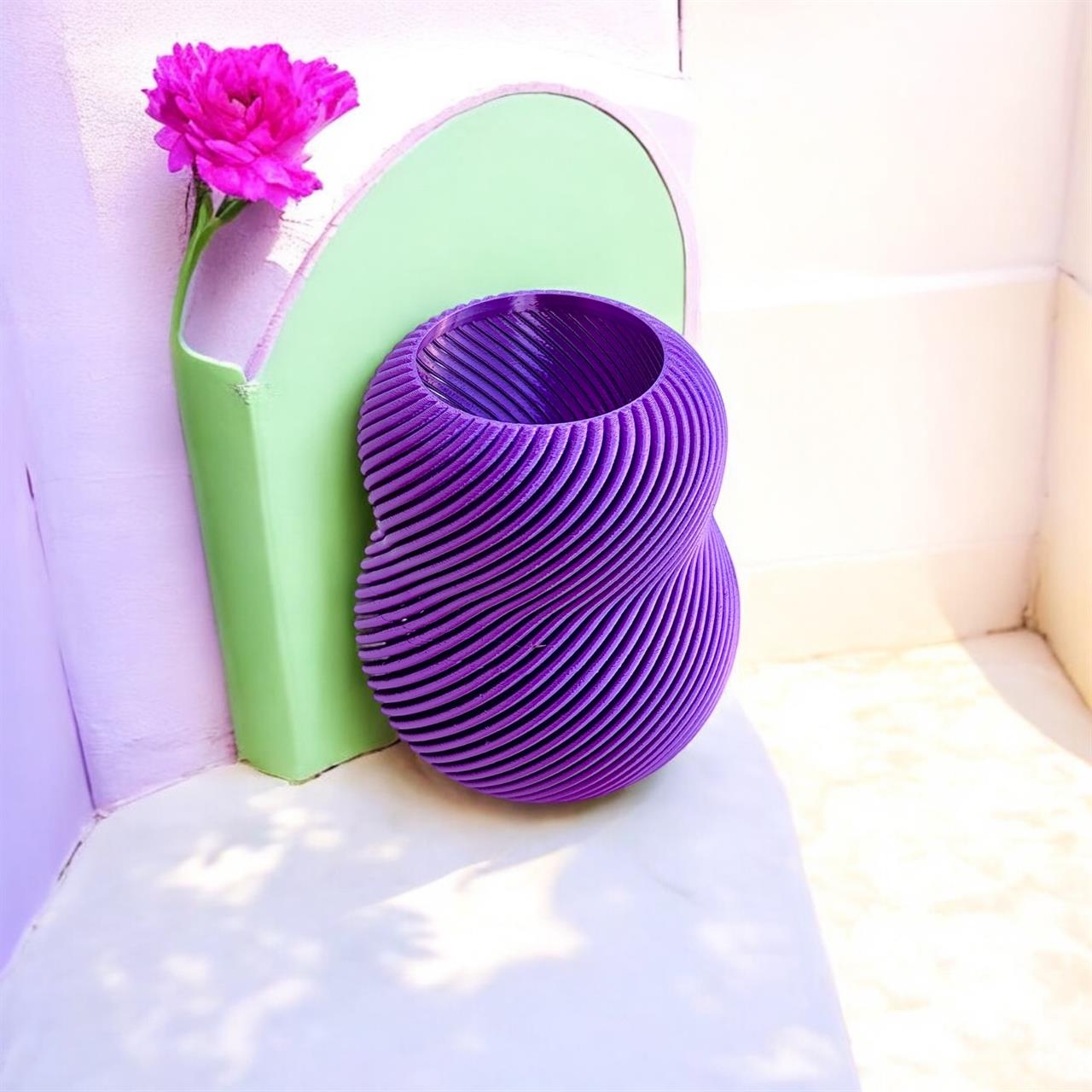 Das Bild zeigt eine violette Vase mit einem modernen, gedrehten Design. Neben der Vase steht eine grüne Dekoration mit einer pinken Blume.