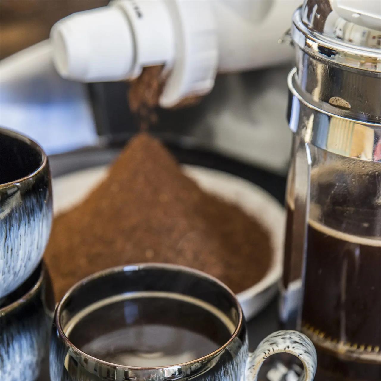 Auf dem Bild ist eine Getreide- und Kaffeemühle zu sehen, aus der frisch gemahlenes Kaffeepulver fällt. Daneben stehen eine Tasse Kaffee und eine Kanne mit frisch gebrühtem Kaffee.