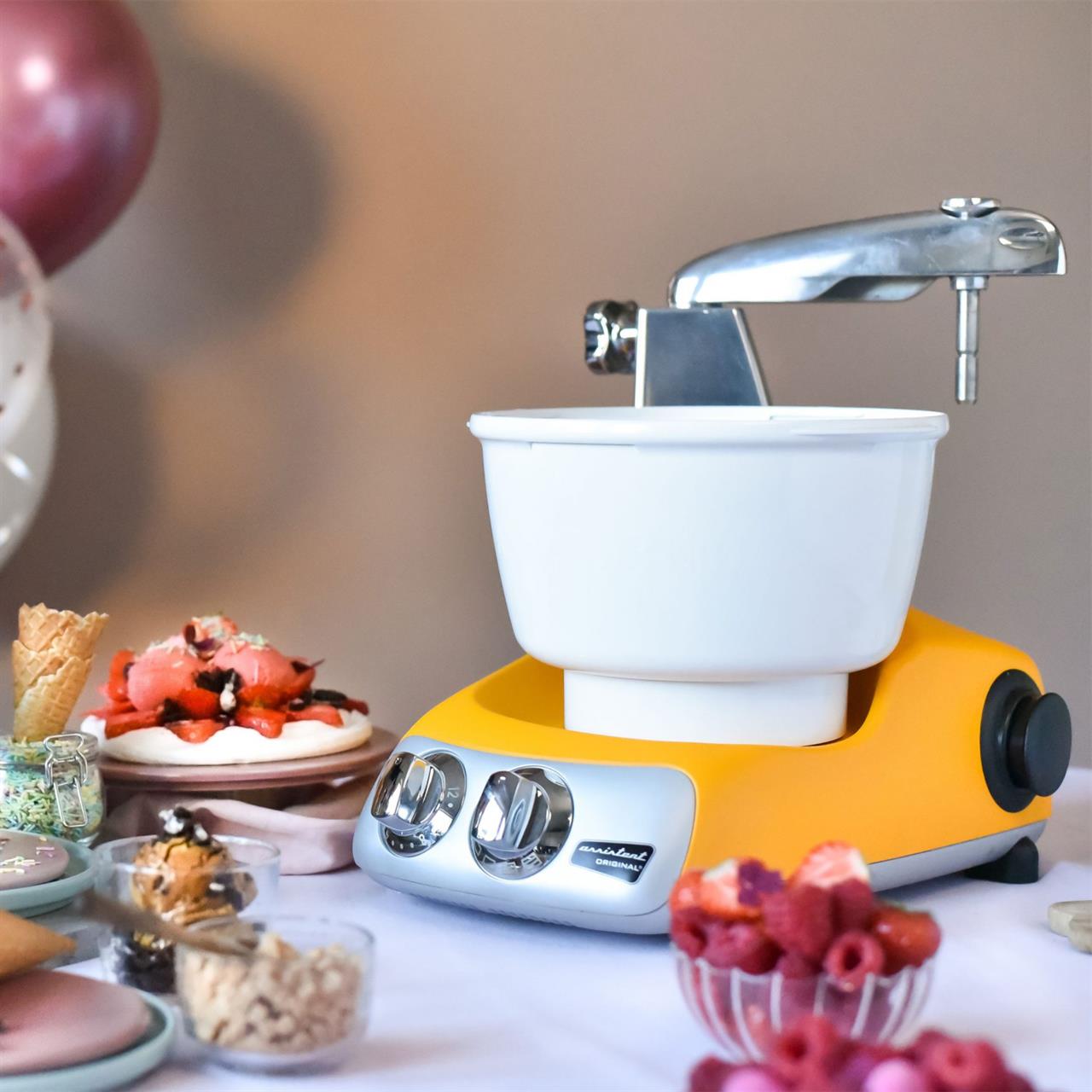 Das Bild zeigt eine gelbe Eismaschine Ankarsrum auf einem Tisch. Rundherum stehen verschiedene Zutaten und Dessertschalen.