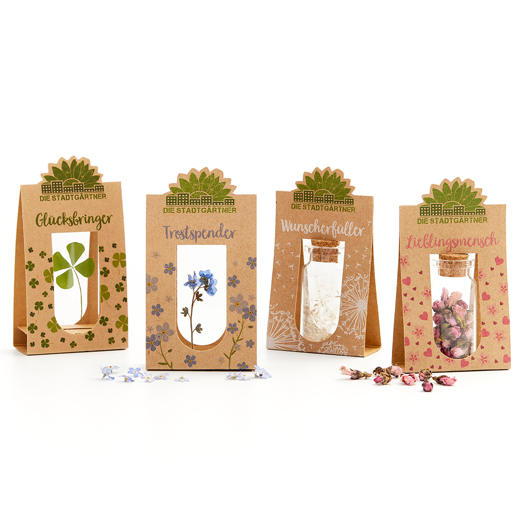 Auf dem Bild sind vier kleine Geschenktüten mit verschiedenen Inhalten zu sehen. Das Produkt "Lieblingsmensch, Emotionsbringer" enthält rosa Blüten in einem kleinen Glas mit Korken.