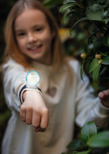Ein Mädchen steht draußen zwischen grünen Pflanzen und lächelt. Sie zeigt stolz ein leuchtendes Armband an ihrem Handgelenk.