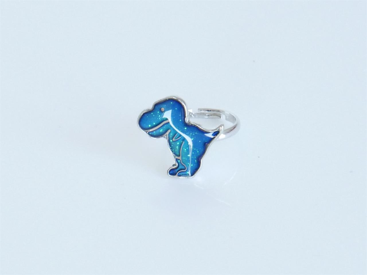 Der Ring zeigt eine blaue Dinosaurierfigur mit glitzernden Akzenten. Er hat eine silberfarbene Fassung und ist verstellbar.