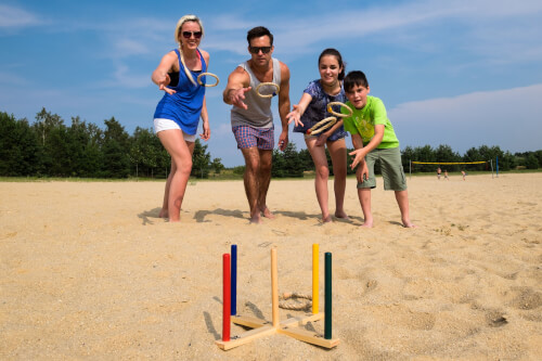 Eine Familie spielt Wurfspiel am Strand. Alle vier werfen Ringe auf bunte Stäbe, die im Sand stehen.