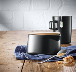 Auf dem Bild ist eine schwarze Zuckerdose namens „Zuckerdose Kineo“ mit Holzdeckel zu sehen. Im Hintergrund steht eine schwarze Tasse mit einem Teebeutel auf einem Holztisch.