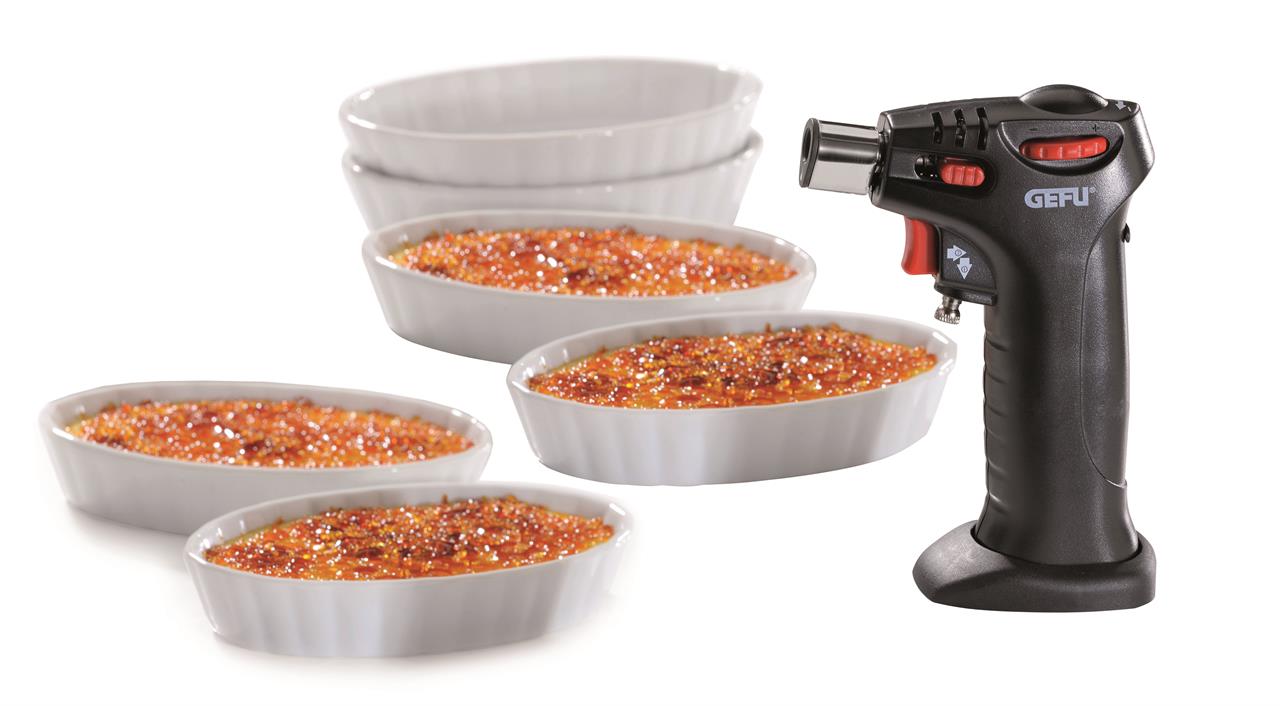 Das Bild zeigt das Crème Brûlée-Set Carmello mit sieben Teilen, darunter vier weiße Förmchen und ein schwarzer Flambierbrenner. In den Förmchen ist fertige Crème Brûlée mit goldbrauner Karamellkruste zu sehen.