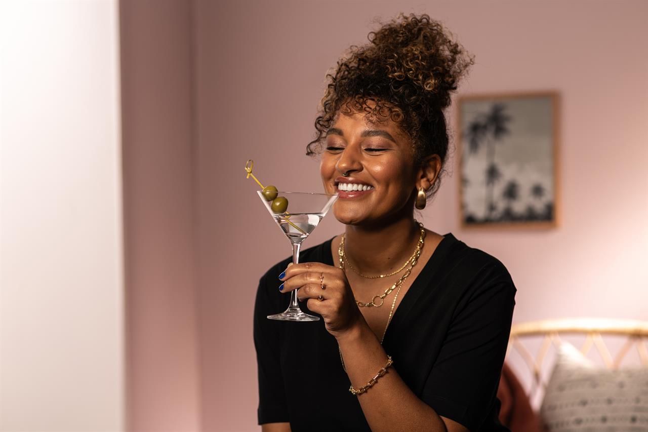 Eine Frau hält ein Martini-Glas mit Oliven und lächelt glücklich. Das Produkt "4er Martini Bar Special 86" wird dabei präsentiert.