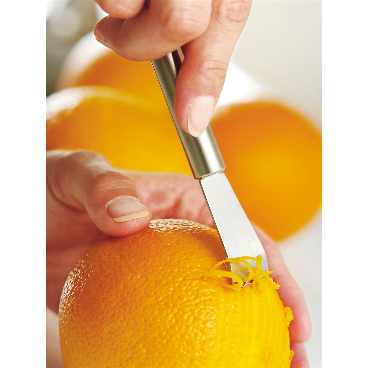 Eine Hand benutzt den Zitronenschaber Profi Plus, um die Schale einer Orange abzuschaben. Mit dem Edelstahlwerkzeug werden feine Zesten von der Frucht gelöst.