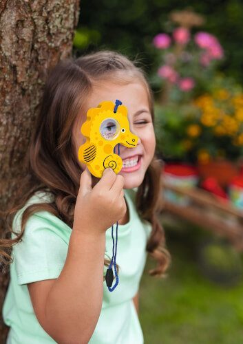 Ein Mädchen steht draußen neben einem Baum und lächelt. Sie hält ein gelbes Spielzeug in Seepferdchenform vor ihr Auge.