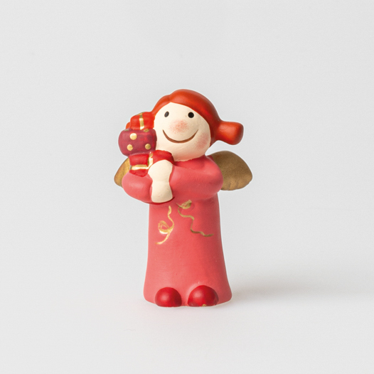 Die Figur zeigt einen Engel mit roten Haaren und goldenen Flügeln. Der Engel trägt ein rotes Kleid und hält ein Geschenk in den Händen.