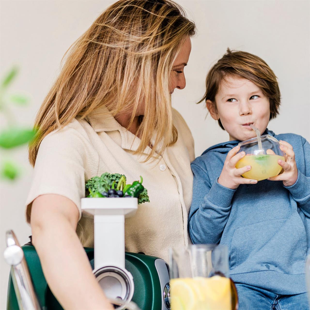 Eine Frau und ein Kind genießen zusammen einen gesunden Saft. Neben ihnen steht das Produkt "Go Green Package" mit frischem Gemüse und Obst.