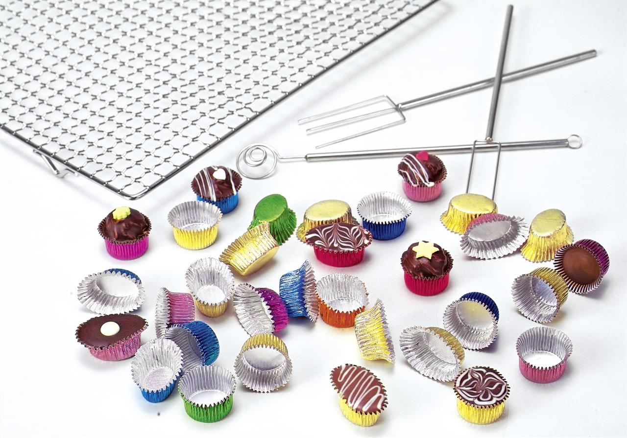 Das Bild zeigt ein Pralinenset mit bunten Förmchen, Pralinen, einem Gitter und Pralinenwerkzeugen. Die Pralinen sind verschieden dekoriert und liegen neben den leeren Förmchen verstreut.