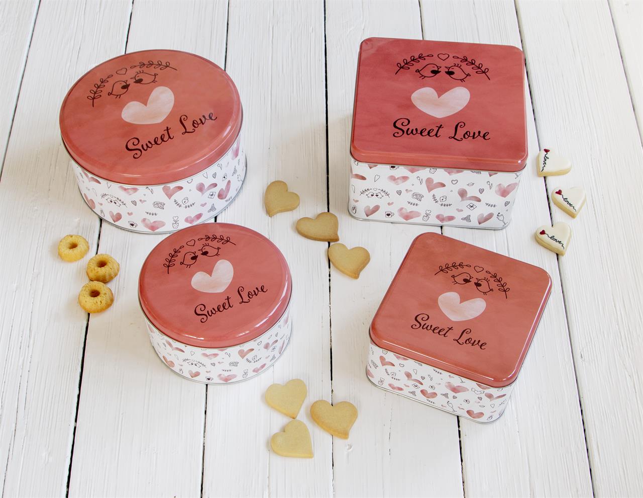 Auf dem Bild sind vier Gebäckdosen mit der Aufschrift "Sweet Love" in rosa und weiß abgebildet. Um die Dosen herum liegen kleine Kekse in Herz- und Ringform auf einem weißen Holztisch.